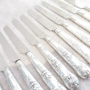 Puede incluir: Un juego de 12 cuchillos de mantequilla plateados con mangos ornamentados. Los cuchillos tienen un acabado pulido y están dispuestos en fila sobre una superficie blanca.