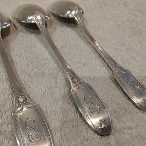 May include: Three silver-toned metal spoons featuring ornate embossed detailing on the handles. Each handle displays a decorative border with leaf motifs and a central engraved script letter S. The spoons have polished, rounded bowls and long stems.