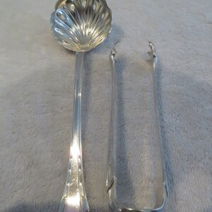 May include: A set of two silver serving utensils. The first utensil is a spoon with a decorative, scalloped bowl and a long, slender handle. The second utensil is a pair of tongs with a rounded, curved end.