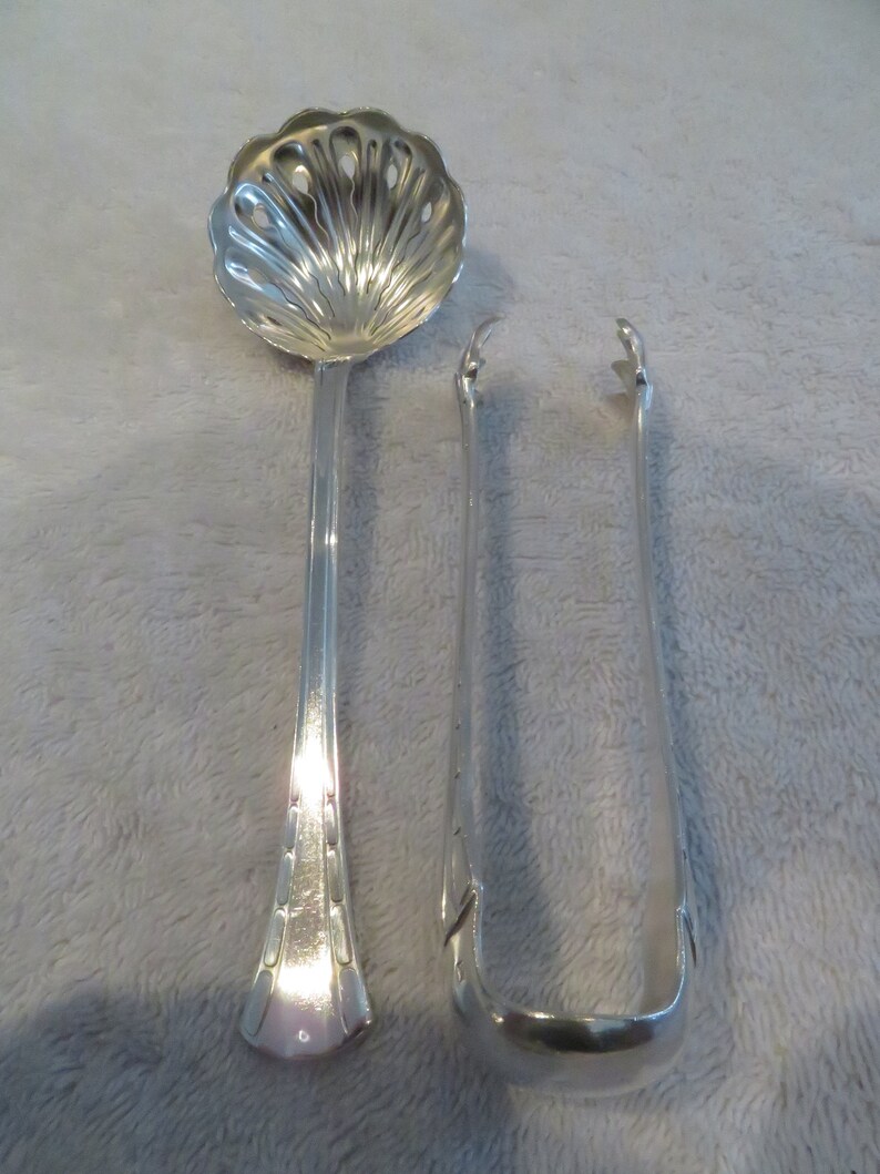 May include: A set of two silver serving utensils. The first utensil is a spoon with a decorative, scalloped bowl and a long, slender handle. The second utensil is a pair of tongs with a rounded, curved end.