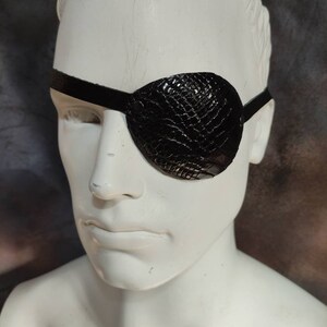 May include: A black leather eye patch with a textured, snake-like pattern. The patch is worn over the right eye and secured with a black strap.
