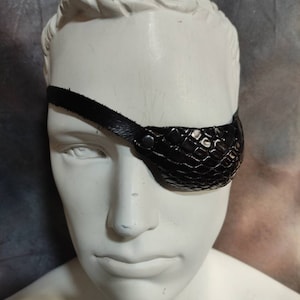 May include: A black leather eye patch with a textured surface. The patch is worn over the right eye and has a strap that goes around the head.