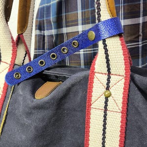 May include: A navy blue canvas tote bag with cream-colored straps featuring red and black stripes. The bag has brown leather accents and a blue strap with metal eyelets and a button closure. The bag is designed for carrying items.