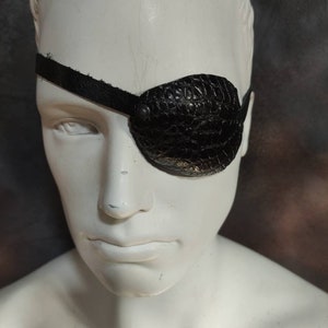 Könnte beinhalten: Ein schwarzes Leder-Augenpflaster mit einer strukturierten Oberfläche und einem schwarzen Band. Das Augenpflaster wird auf einem weißen Mannequin-Kopf getragen.