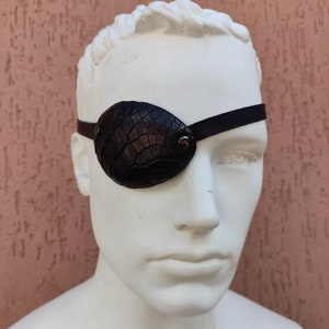 Może przedstawiać: Czarna skórzana opaska na oko z krokodylim wzorem. Opaska jest noszona na białej głowie manekina.