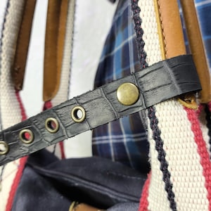 May include: A blue canvas tote bag with red, white, and blue striped straps. The bag has a black leather strap with metal eyelets and a brass buckle. The bag is made of a durable canvas material and has a spacious interior.