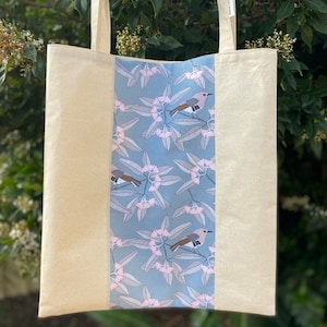 May include: A white canvas tote bag with a blue and pink floral print featuring birds. The bag has two long handles.