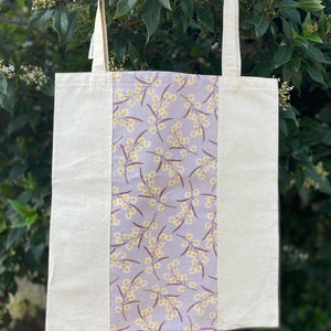 May include: A white tote bag with a grey and yellow floral print panel. The bag has two long straps.