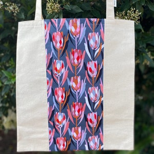 May include: A beige tote bag with a gray and pink floral print panel. The print features stylized flowers with red, pink, orange, and blue accents. The bag has a label that reads "Simply Handmade".