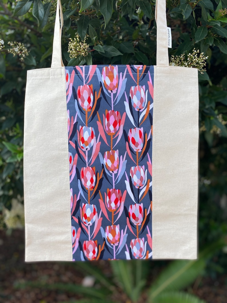 May include: A beige tote bag with a gray and pink floral print panel. The print features stylized flowers with red, pink, orange, and blue accents. The bag has a label that reads "Simply Handmade".