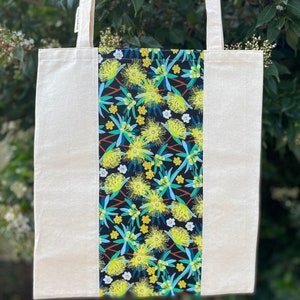 May include: A natural canvas tote bag with a black and yellow floral print panel. The print features yellow flowers with green leaves on a black background.