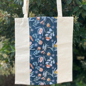 May include: A beige tote bag with a dark blue fabric panel featuring a floral pattern of white, pink, and yellow flowers and leaves. The bag has two long beige straps.