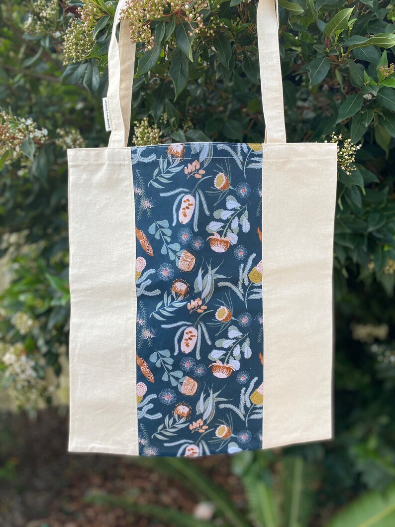 May include: A beige tote bag with a dark blue fabric panel featuring a floral pattern of white, pink, and yellow flowers and leaves. The bag has two long beige straps.