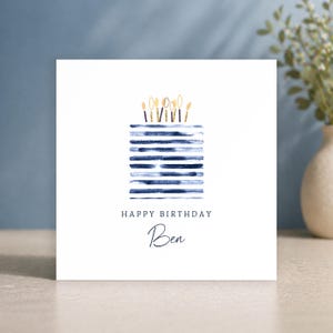 Puede incluir: Una tarjeta de felicitaci&oacute;n cuadrada con fondo blanco que presenta una ilustraci&oacute;n en acuarela de un pastel hecho de rayas horizontales azul marino. Encima hay velas con purpurina dorada. El texto dice HAPPY BIRTHDAY y el nombre Ben.
