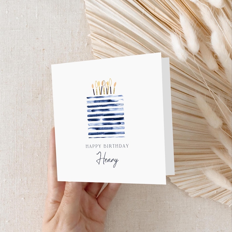 Puede incluir: Una tarjeta de felicitaci&oacute;n cuadrada de color blanco con una ilustraci&oacute;n en acuarela de un pastel con rayas azules y velas doradas. El texto dice HAPPY BIRTHDAY seguido del nombre Henry en una fuente cursiva negra bajo el pastel.