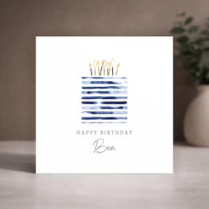 Puede incluir: Una tarjeta de felicitaci&oacute;n cuadrada de color blanco con una ilustraci&oacute;n en acuarela de un pastel hecho de rayas horizontales azul marino coronado con velas de purpurina dorada. El texto dice HAPPY BIRTHDAY y el nombre Ben en cursiva.