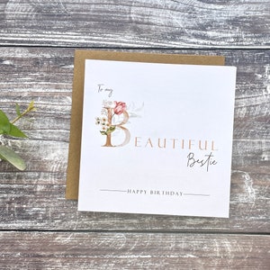 Pu&ograve; includere: Un biglietto bianco con un disegno floreale che presenta una lettera dorata "B" e il testo "To my beautiful bestie - Happy Birthday".