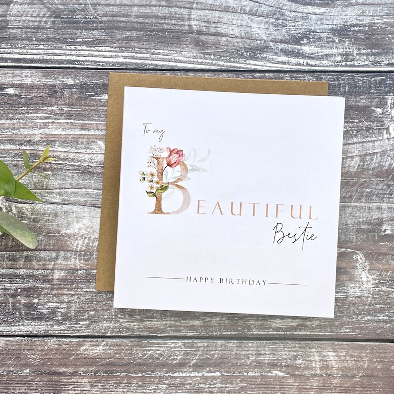 Pu&ograve; includere: Un biglietto bianco con un disegno floreale che presenta una lettera dorata "B" e il testo "To my beautiful bestie - Happy Birthday".
