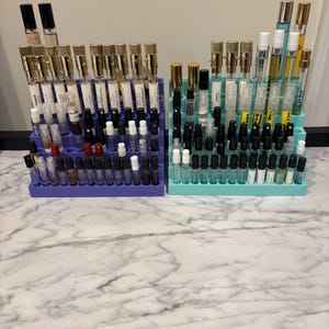 Peut inclure: Deux présentoirs à échantillons de parfum, un violet et un turquoise, chacun rempli de nombreux petits flacons de parfum. Les flacons ont différentes couleurs de bouchons, dont l'or, le noir et l'argent. Les présentoirs sont sur une surface à motifs de marbre.