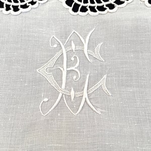 May include: White embroidered linen fabric with a delicate lace trim and a monogram in the center. The monogram is a stylized "E" with a flourish.