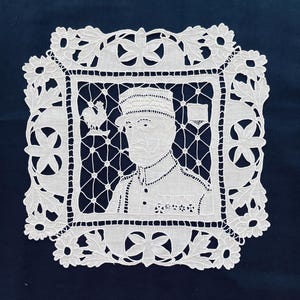 May include: White lace doily featuring a portrait of a man in a military uniform, framed by a floral and geometric design. The portrait is detailed, with a rooster emblem visible. The doily is set against a dark blue background.