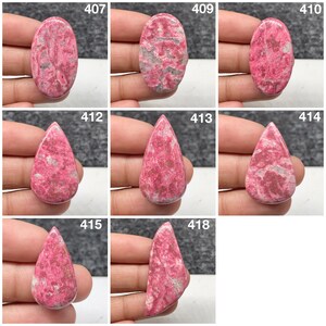 May include: A collection of eight polished rhodonite cabochons labeled 407, 409, 410, 412, 413, 414, 415, and 418. These stones feature vibrant pink and magenta hues with mottled grey and black inclusions, presented in various oval, teardrop, and asymmetrical shapes.