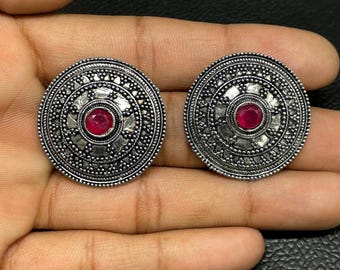 以下が含まれることがあります： 複雑な質感の金属模様と中央に円形のファセットカットされた赤い石をあしらった、銀色の丸いスタッドピアス一対。デザインには、小さな隆起した金属ビーズの同心円と幾何学的なディテールが含まれています。