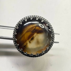 Puede incluir: Un anillo redondo con una piedra de ágata pulida, translúcida y con bandas de color marrón y crema, engastada en un bisel de metal plateado. El engaste tiene un borde decorativo de cuerda trenzada y garras dentadas que sujetan el cabujón.