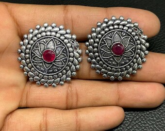 以下が含まれることがあります： 酸化銀色の丸いスタッドピアス一対。中央に円形の赤い石があしらわれています。デザインには詳細な花柄のモチーフが施され、その周囲を小さな隆起した銀色のビーズが円状に囲むテクスチャーのある縁取りが特徴です。
