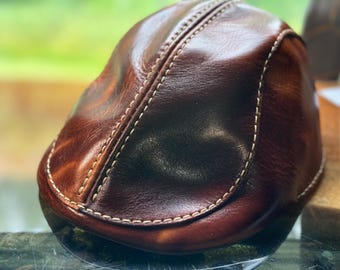 May include: A brown leather flat cap with white stitching. The cap has a smooth, shiny finish.