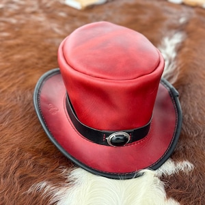 May include: A red leather top hat with a black band and a silver button. The hat is sitting on a brown and white cowhide.