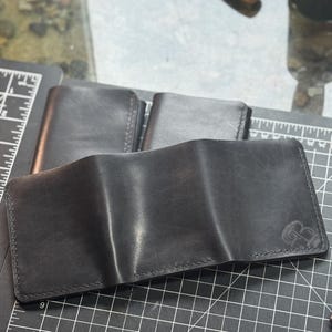 May include: Black leather wallet, open and displayed, with a subtle embossed design in the corner. The wallet is on a black cutting mat with other closed wallets in the background. The leather appears to be smooth and well-crafted.