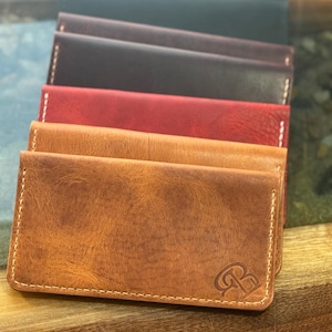 May include: A stack of leather wallets in various colors, including brown, red, and black. The wallets are rectangular with rounded corners and white stitching. The front wallet has a debossed logo in the bottom right corner.