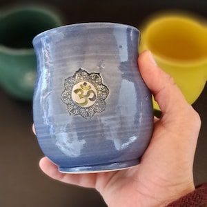Pode incluir: Uma caneca de cerâmica azul com um símbolo Om prateado. A caneca tem formato arredondado e uma borda ligeiramente alargada. O símbolo Om está centrado na frente da caneca. Outras canecas verdes e amarelas estão no fundo.