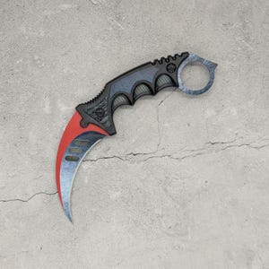 Può includere: Un coltello karambit curvo con manico testurizzato nero e blu e un anello per il dito all'estremità. La lama presenta un design bicolore con una sezione rosso vivo vicino all'impugnatura e una finitura blu metallizzato sul filo curvo.