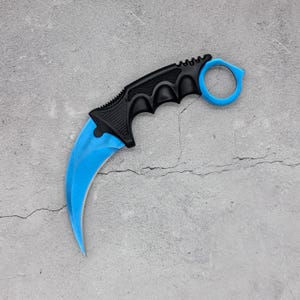 May include: A karambit knife featuring a curved, bright blue blade and a black handle with a textured grip. The handle includes a prominent finger ring at the end, designed for a secure hold. The blade has a sharp, pointed tip and a curved edge.