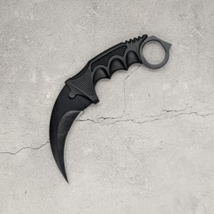 May include: A matte black karambit knife featuring a curved blade and a textured handle with a finger ring at the pommel. The design includes a ridged spine and a grip area with moulded finger grooves, showcasing a sleek, solid black finish throughout.