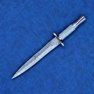 May include: A double-edged dagger featuring a pointed blade with a distinct wavy, layered metal pattern. The handle is made of polished silver-toned metal with a textured, cross-hatched grip and a small, straight crossguard separating it from the blade.