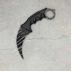 Könnte beinhalten: Ein gebogenes Karambit-Messer mit mattschwarzem Finish und beigefarbenem Tigerstreifenmuster. Der Griff verfügt über einen Fingerring am Ende und einen strukturierten Griffbereich. Die Klinge ist stark gekrümmt und folgt der Sichelform.