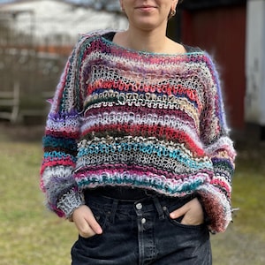 Könnte beinhalten: Ein bunter gestreifter Pullover mit lockerer Passform und leicht verkürzter Länge. Der Pullover ist aus weichem, flauschigem Garn gefertigt und hat einen lässigen, entspannten Stil.