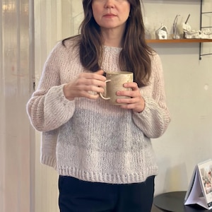 Peut inclure: Un pull en tricot rose clair, gris et blanc, avec un col rond et des manches longues. Le pull est porté avec un pantalon noir. Une tasse dorée est tenue.