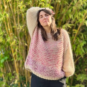 Può includere: Un maglione lavorato a mano a maglia con una sfumatura di colore dal beige chiaro al rosa e al viola. Il maglione ha uno scollo tondo e maniche lunghe e oversize. Sullo sfondo, steli di bambù verdi.
