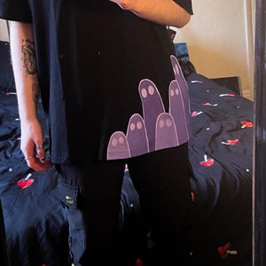 May include: Black t-shirt featuring a purple ghost design. The outfit includes black cargo trousers. A silver chain necklace and a phone with a space-themed case are also visible.