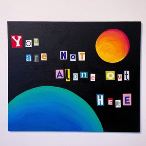 May include: A black canvas with the words "You are Not Alone Out Here" spelled out in colourful letters. A blue planet and a yellow sun are painted in the background.