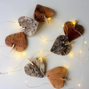 May include: A string of fairy lights with wooden heart-shaped ornaments. The hearts are made of natural birch bark and have a rustic, natural look. The lights are strung together with small, clear bulbs.