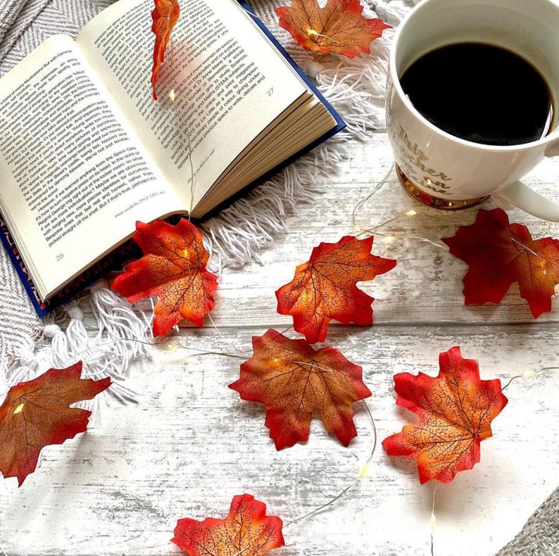 May include: A white wooden surface with a book, a cup of coffee, and a string of fairy lights shaped like fall leaves. The leaves are orange and red.