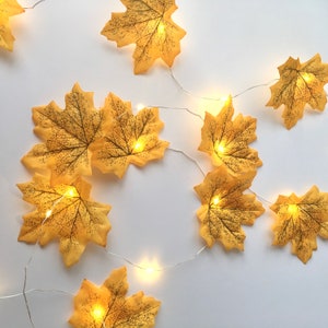 May include: A string of fairy lights with yellow maple leaf cutouts. The lights are strung on a silver wire and the leaves are evenly spaced.
