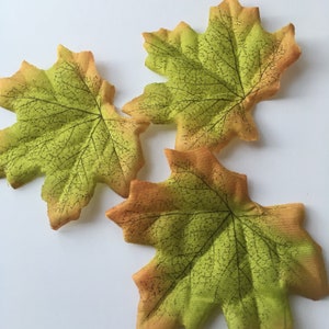 May include: Three artificial maple leaves in shades of green and orange. The leaves have a detailed vein pattern and a slightly textured surface.