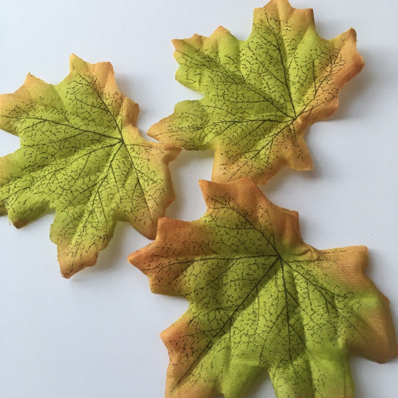 May include: Three artificial maple leaves in shades of green and orange. The leaves have a detailed vein pattern and a slightly textured surface.