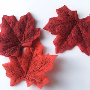 May include: Three red fabric maple leaves with black veins. The leaves are arranged in a cluster on a white background.